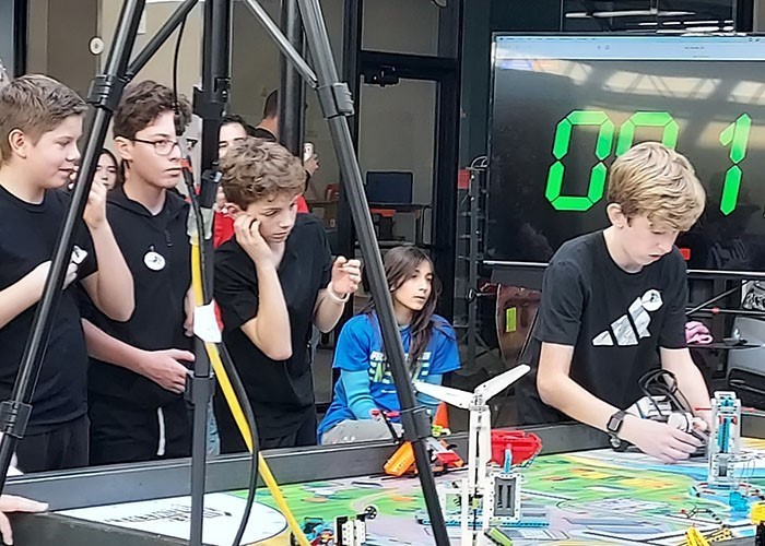 Image of 4 young teenaged boys standing at a work table with one boy working on a LEGO robot with a competition clock in the background