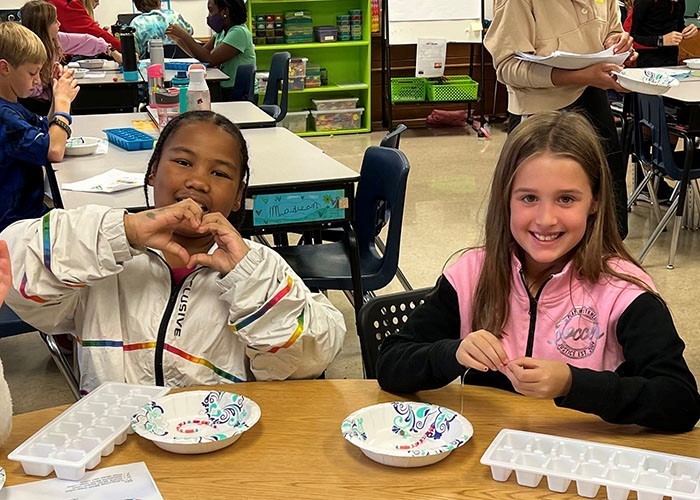 Image of 2 young students at a table with the girl on the left making a heart with her hands while looking at the camera and the girl on the right looking at the camera
