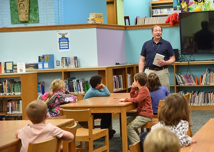Image of a male librarian standing in a school library talking to students who are sitting at tables facing him