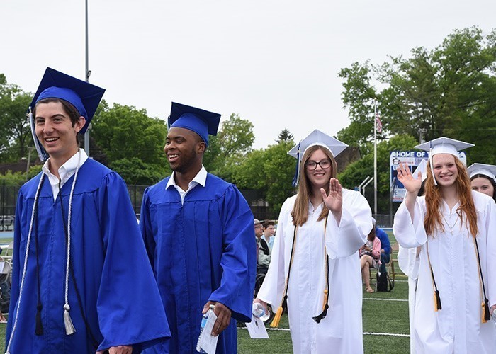 Image of 4 graduates in cap and gown with two male high school graduates in royal blue cap and gown on the left parading in during graduation and two females in white cap and gown waving to the camera on the right