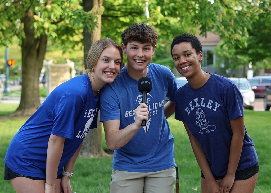 Image of a high school boy holding a microphone and standing between two high school girls, smiling at the camera