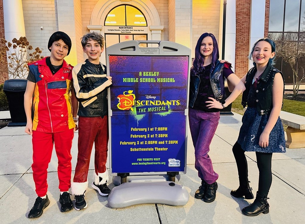 Image of 4 middle school students, 2 boys on the left and 2 girls on the right, who are in costume and standing next to a Disney Descendants The Music promotional board
